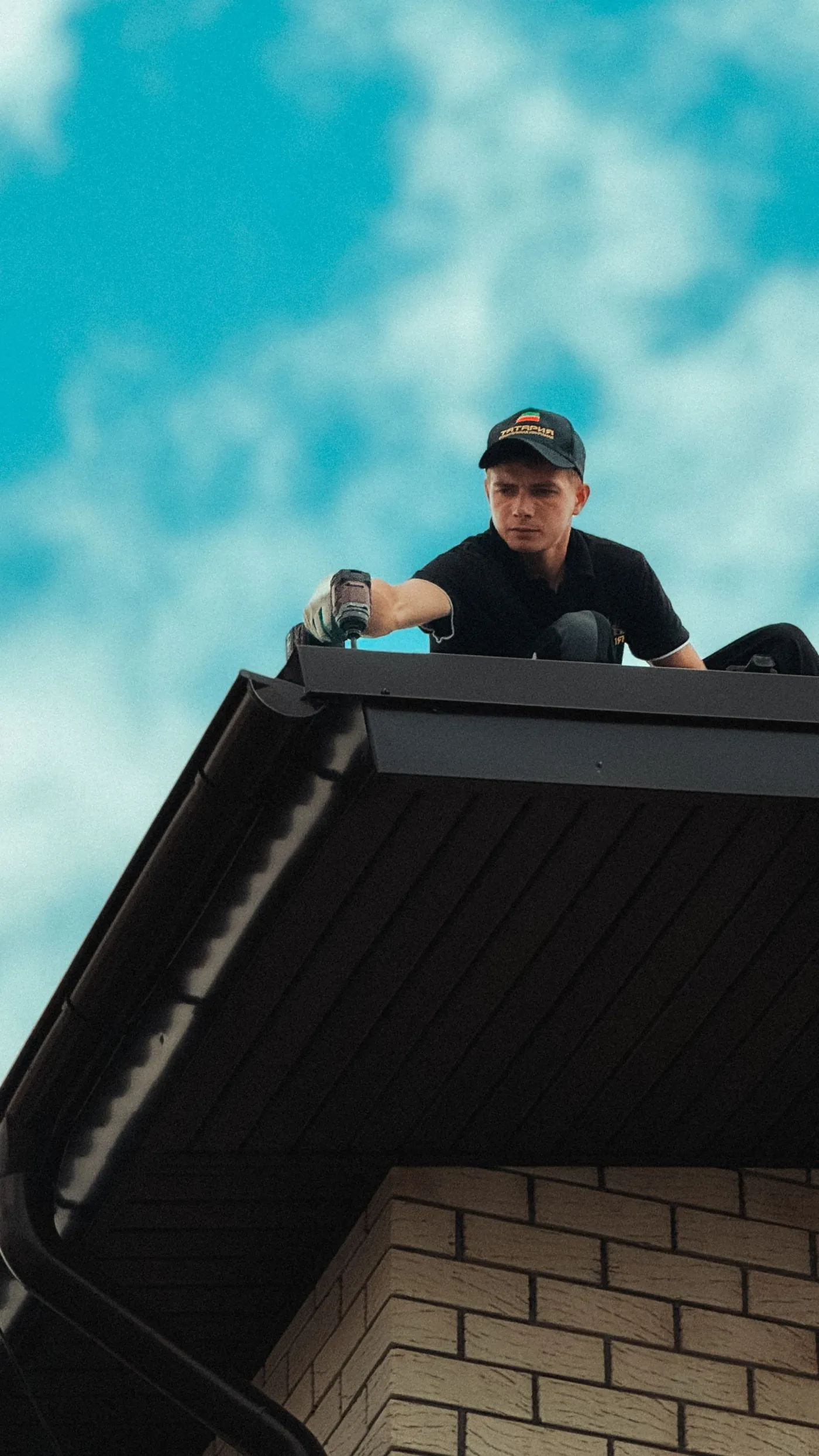 An installer driving a screw at the edge of a dark metal roof eave above a black gutter run and a brick wall