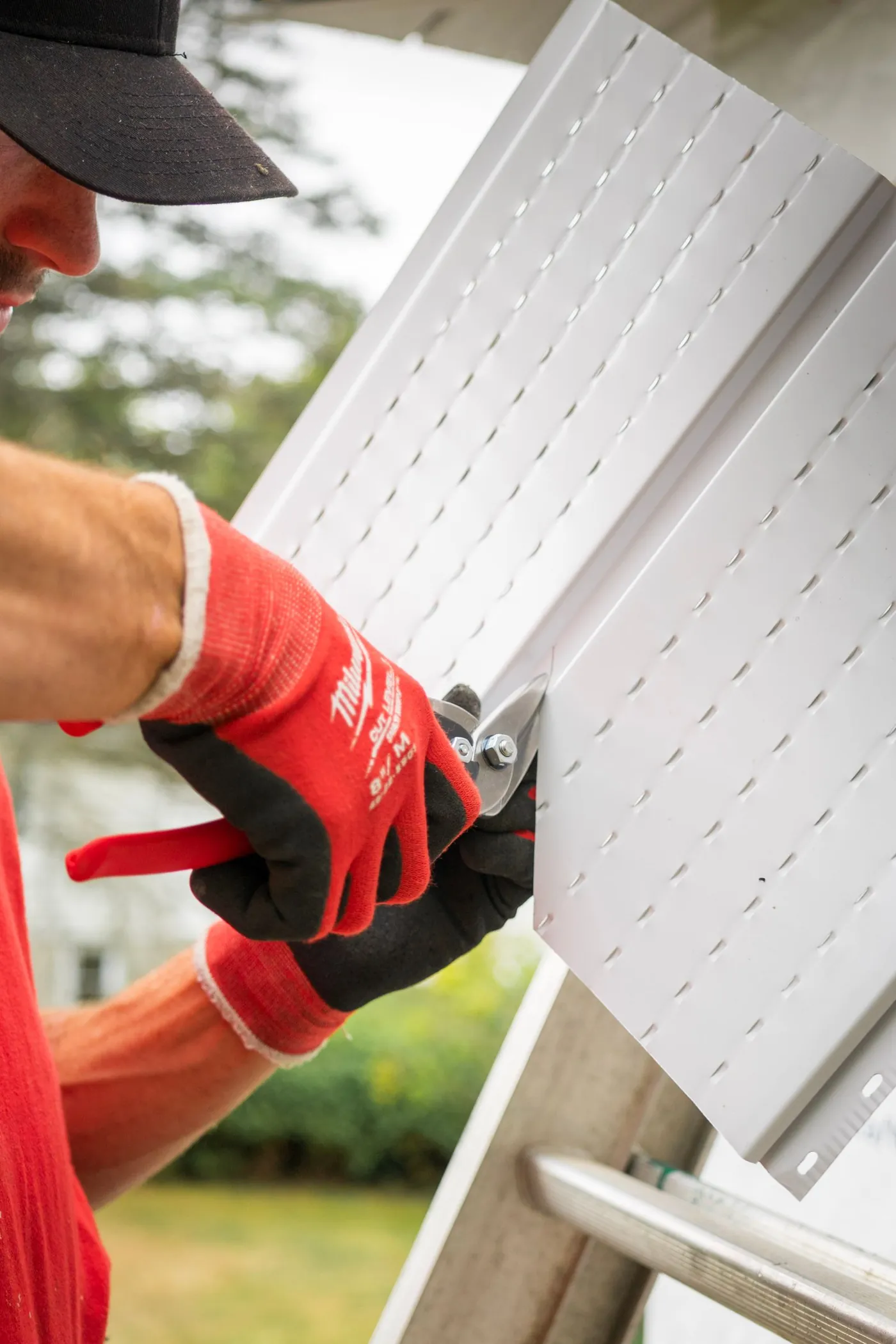 Gloved hands trimming a white vented vinyl panel with tin snips beside a ladder